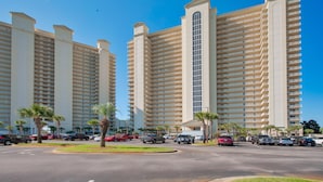 A high-rise hotel with multiple floors, a central elevator, and a parking lot with several cars. - Modern Gulf-View Reno! Steps to Pool, Easy walk to Beach + Beach Gear & Wagon (Miramar Beach, Destin)