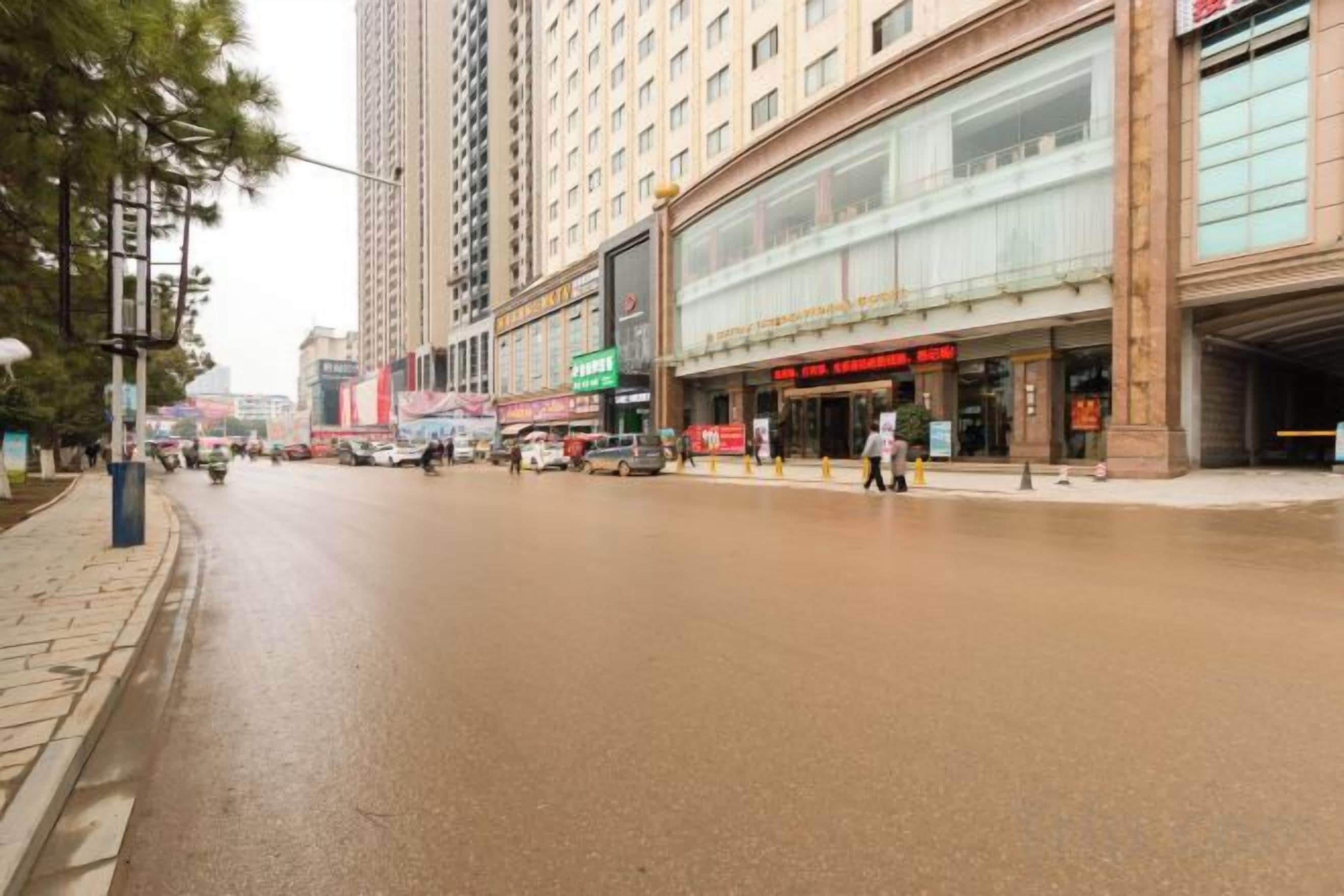 A street with a wet road, tall buildings, and a large glass-fronted building with signage.