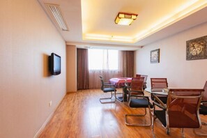 Hotellihuone, jossa on puulattia, seinälle asennettu televisio, ruokapöytä tuoleineen ja ikkuna, jossa on verhot.