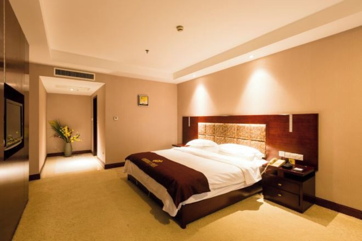 A hotel room with a large bed, a bedside table with a lamp, a wall-mounted TV, and a small plant.