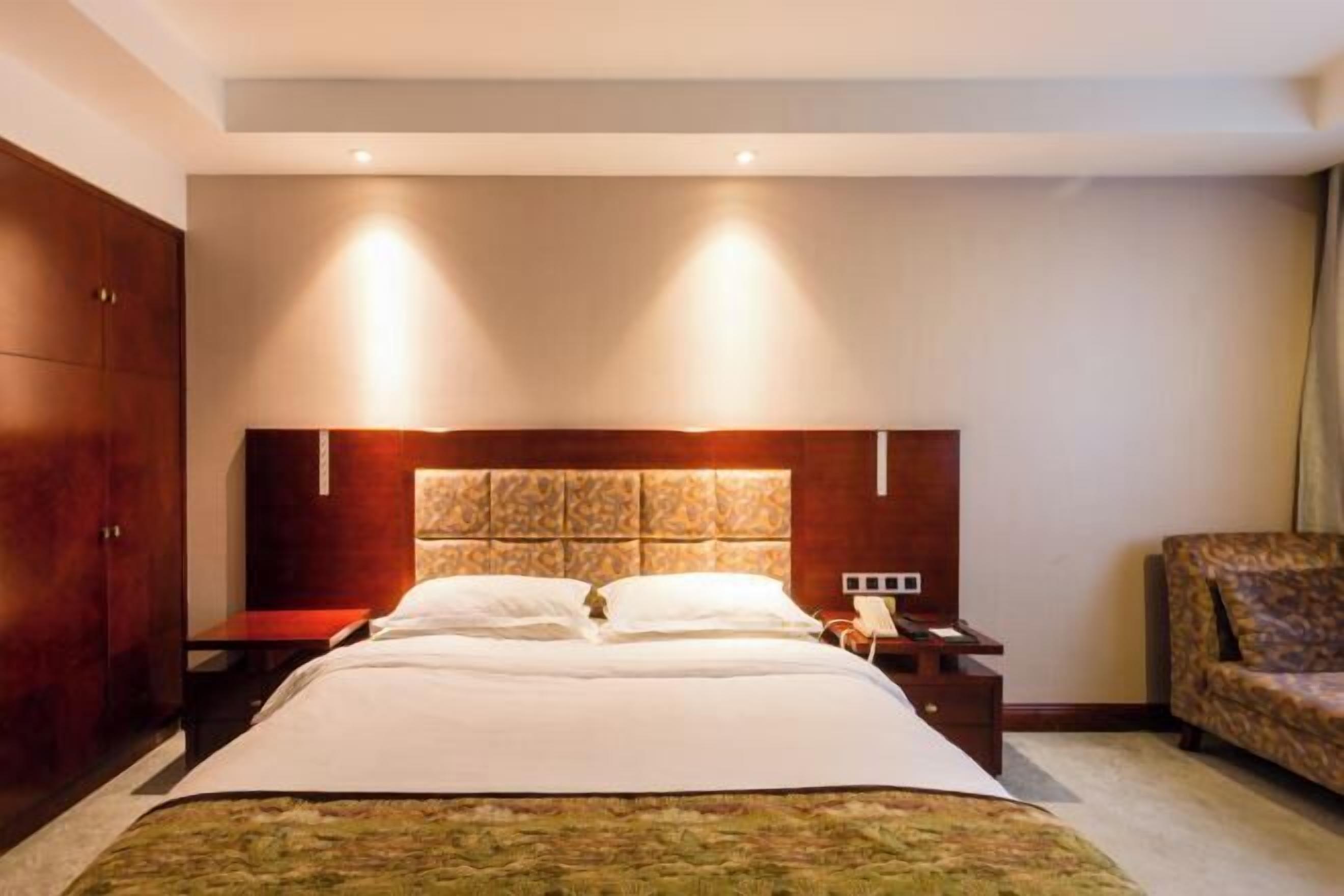 Una camera d'albergo con un letto grande, una testiera in legno, un comodino, un telefono e una poltrona con motivo decorativo.