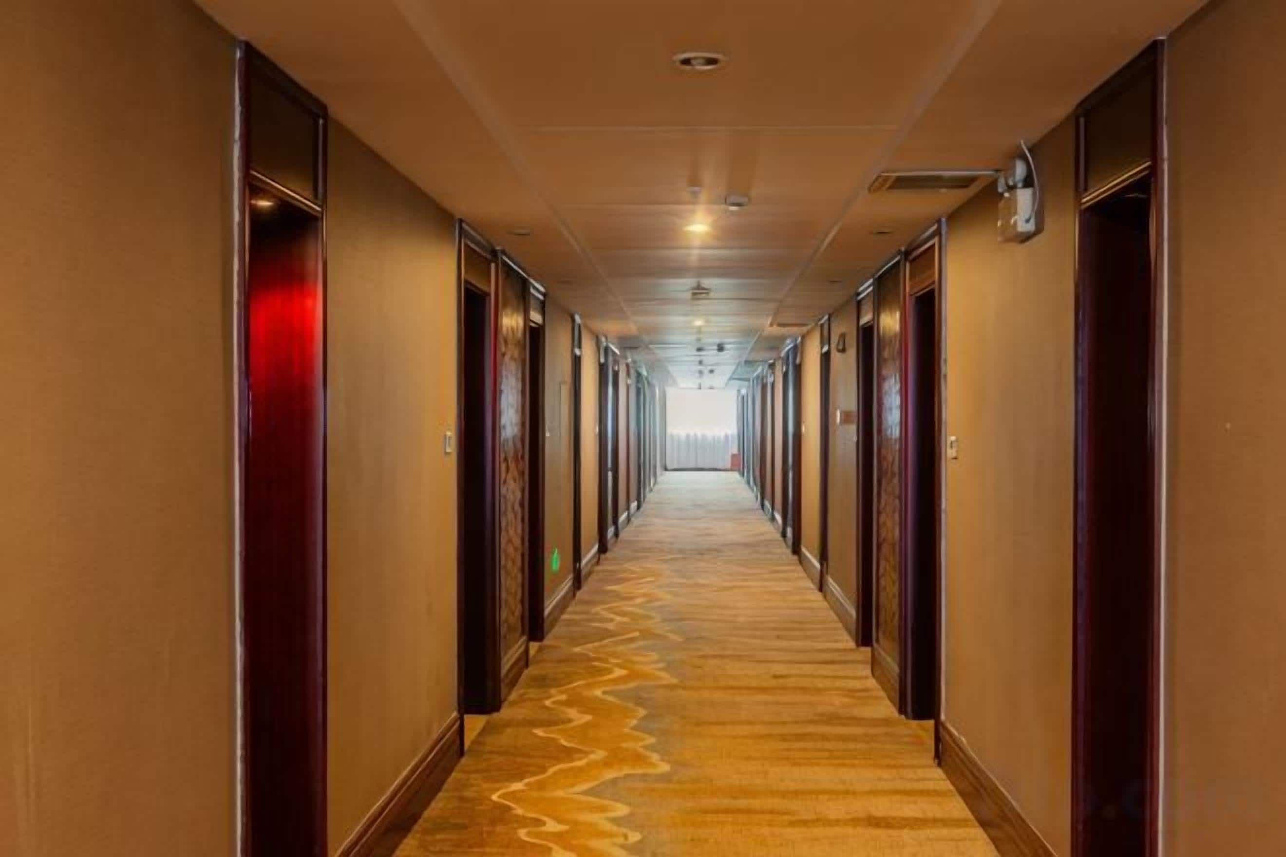 A long, narrow hotel corridor with doors on both sides, carpeted floors, and a ceiling with recessed lighting.