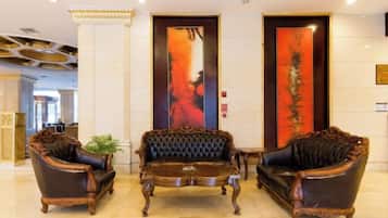 A lobby with three ornate armchairs and a central coffee table, a large vertical painting on the wall, and a reception desk in the background.