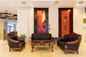 Un vestíbulo con tres sillones decorativos y una mesa de centro, un cuadro vertical grande en la pared, y un mostrador de recepción al fondo.