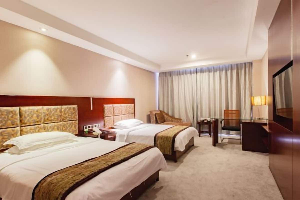 Habitación de hotel con dos camas, un escritorio, una silla, un televisor y un ventanal grande con cortinas.