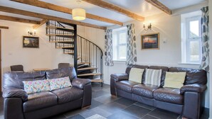 A spacious living room with two leather sofas, a wooden staircase, and a large window with patterned curtains. - Bunk House - 4 Bedroom - Llangenith (Swansea)