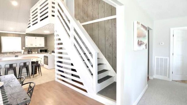 A modern kitchen with a central island, a staircase with white railings, and a dining area with a table and chairs. - Sugarwood Boathouse (Ten Mile)