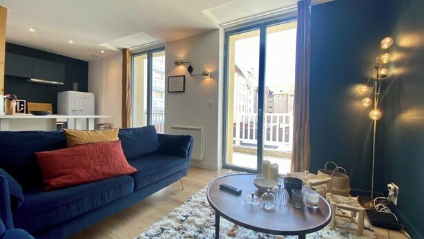 Un salon moderne avec un canapé bleu foncé, une table basse ronde agrémentée d’objets décoratifs, un coin cuisine avec un réfrigérateur blanc et de grandes fenêtres offrant une vue sur la ville.