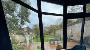 Ein Zimmer mit großem Fenster und Blick auf einen Garten, ein Schachbrett auf einem Tisch und zwei blaue Stühle.