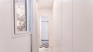 Um corredor estreito com paredes brancas e chão xadrez, dando acesso a uma porta.