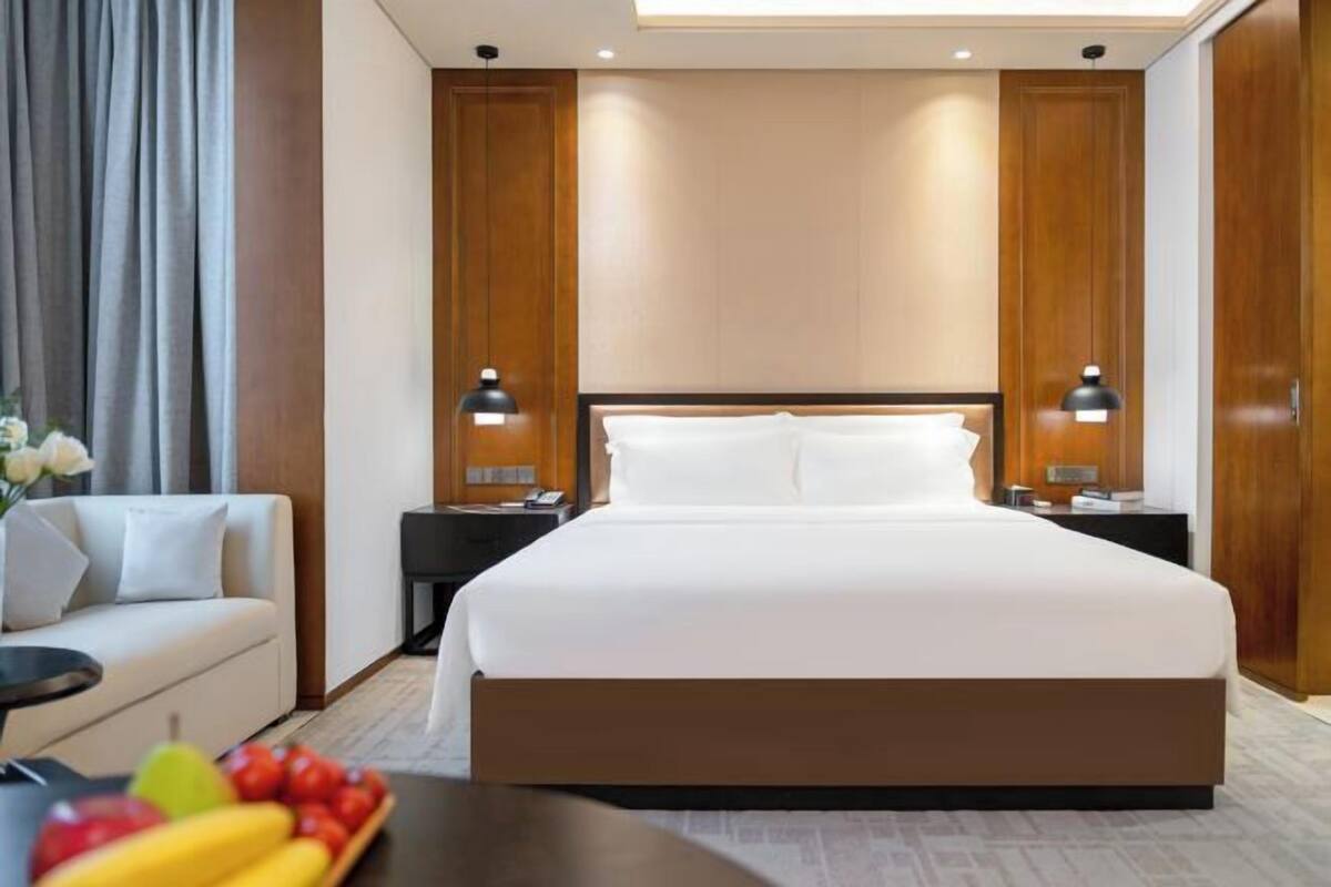 A modern hotel room with a large bed, two bedside tables, a sofa, and a fruit bowl.
