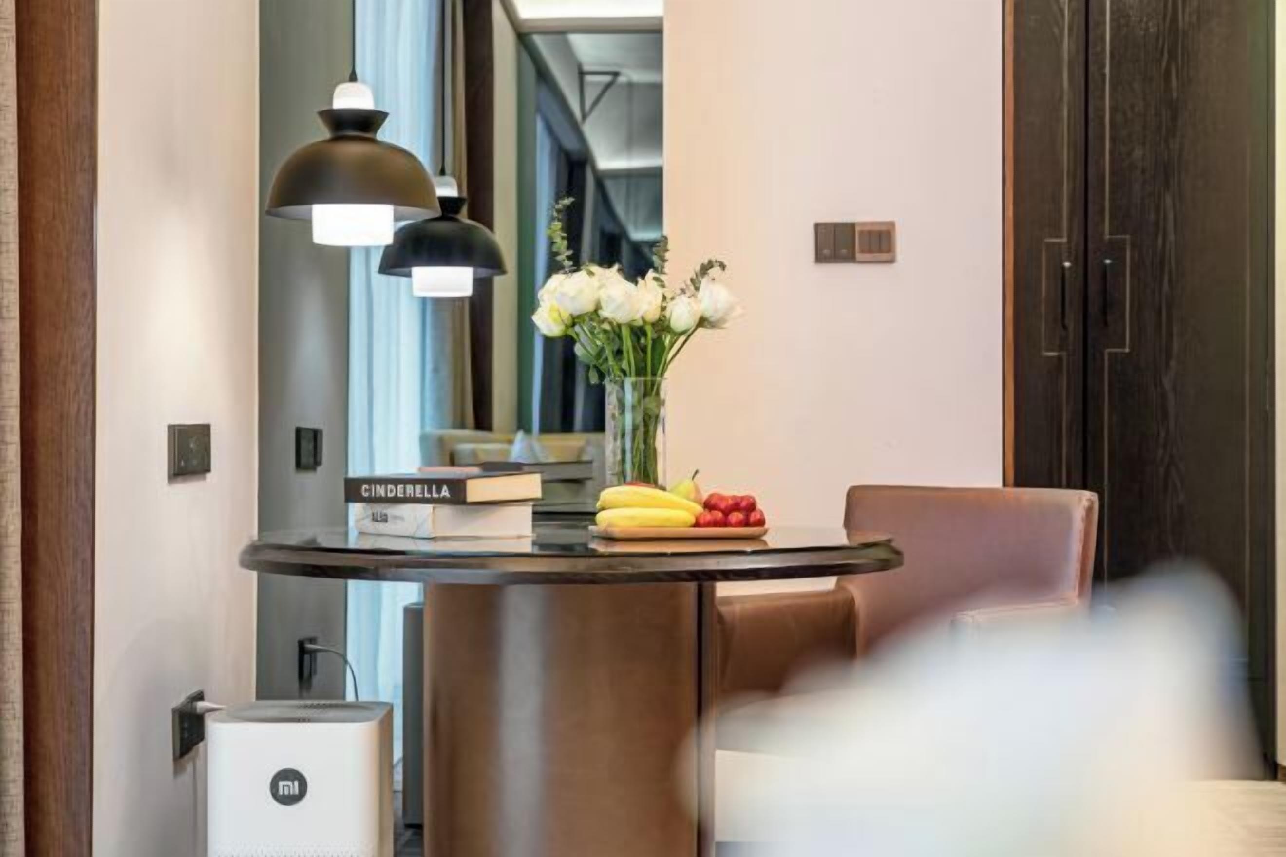 A modern hotel lobby with a round table, a black lamp, a vase with white flowers, and a bookshelf with books.