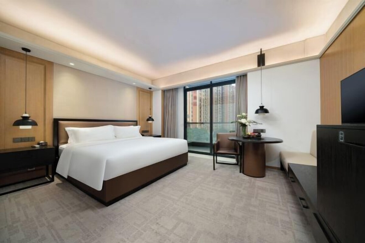 A modern hotel room with a large bed, a desk with a chair, a TV, and a window with curtains.