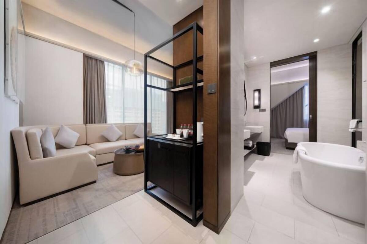 A modern living room with a sofa, a coffee table, and a black cabinet. There is a large window with curtains, a hanging light fixture, and a white bathtub in the adjacent bathroom.