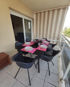 Un balcone con un tavolo da pranzo per quattro persone, circondato da sedie nere, uno sgabello di legno e una porta vetrata che dà su un ambiente buio.