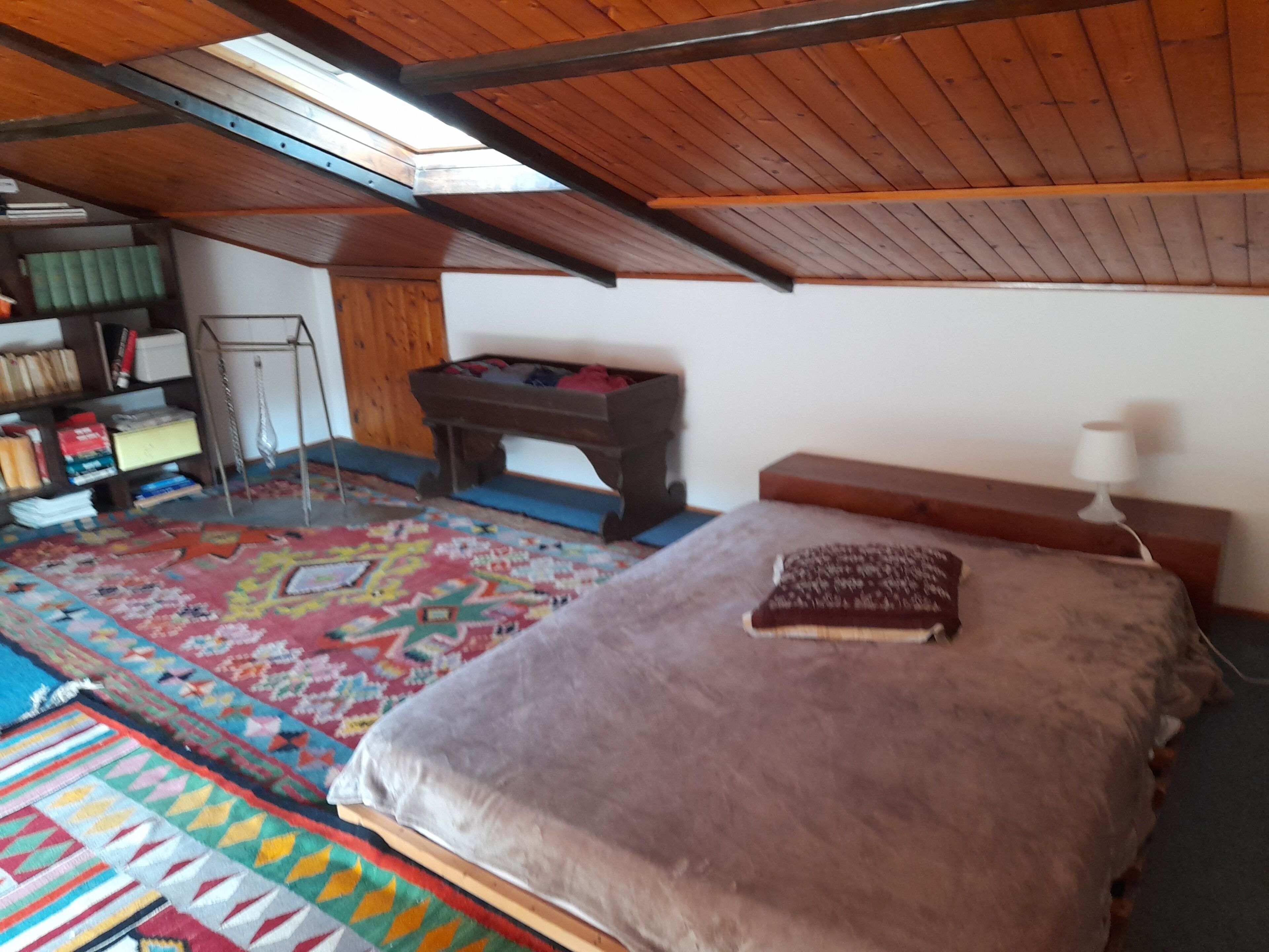 Ein Zimmer mit Hochladerbett, einem kleinen Tisch, einem Bücherregal und einem farbenfrohen Teppich.