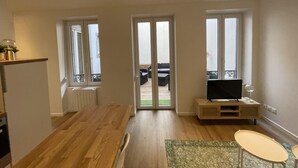 A modern living room with a wooden floor, a TV on a wooden stand, and a view through a glass door to an outdoor area. - Newly renovated apartment, a stone's throw from town center and station (Strasbourg)