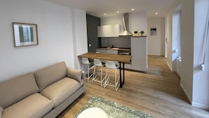 A modern living room with a sofa, a wooden dining table, and a kitchen area in the background. - Newly renovated apartment, a stone's throw from town center and station (Strasbourg)