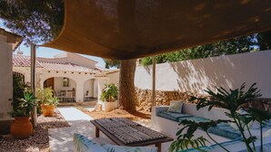 Un patio con mesa de madera, sofá blanco con cojines verdes, plantas en macetas y un muro de piedra.