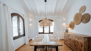 Un comedor con una mesa de madera, sillas blancas, una lámpara de techo trenzada y un mueble de madera con individuales tejidos.