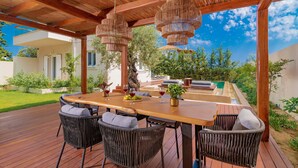 An outdoor dining area with a wooden table and wicker chairs, surrounded by a wooden pergola and hanging wicker chandeliers. - Oresteia Executive Villa, a Flamboyant Retreat (Tragaki)