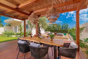 An outdoor dining area with a wooden table and wicker chairs, surrounded by a wooden pergola and hanging wicker chandeliers.