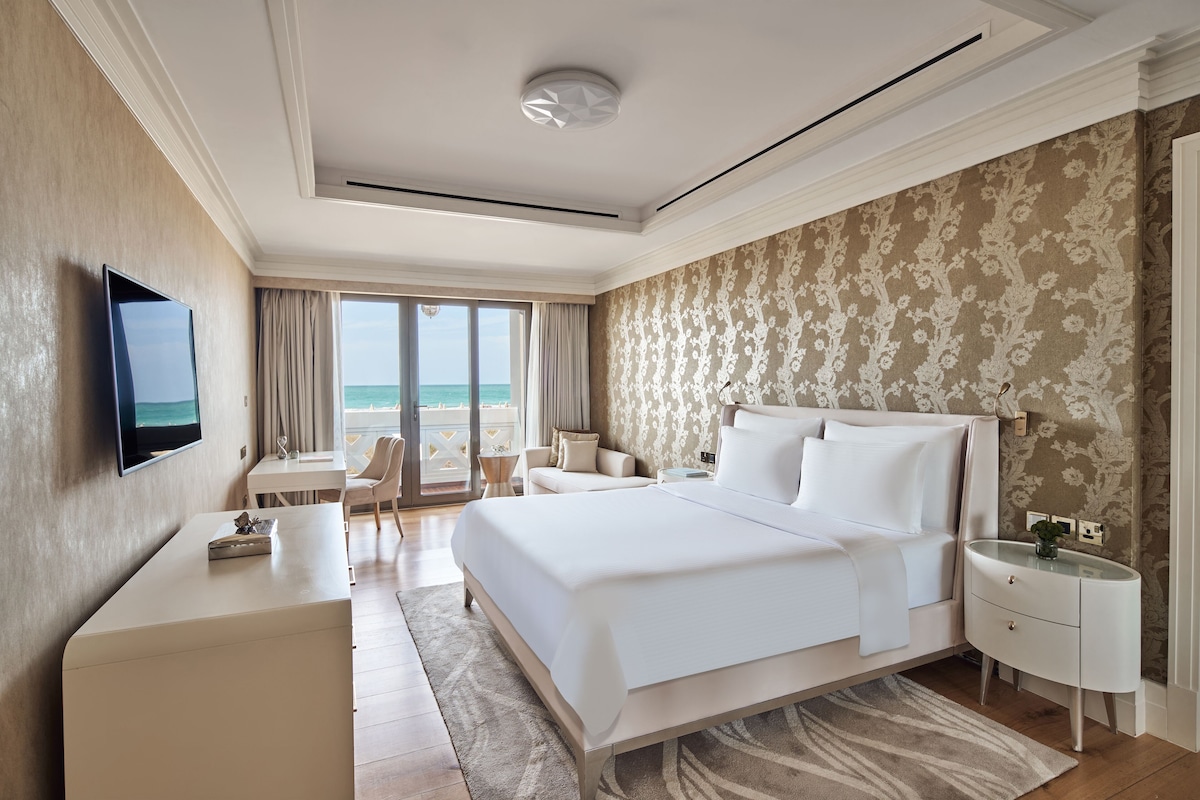 A modern hotel room with a large bed, a TV, a desk, and a view of the sea.