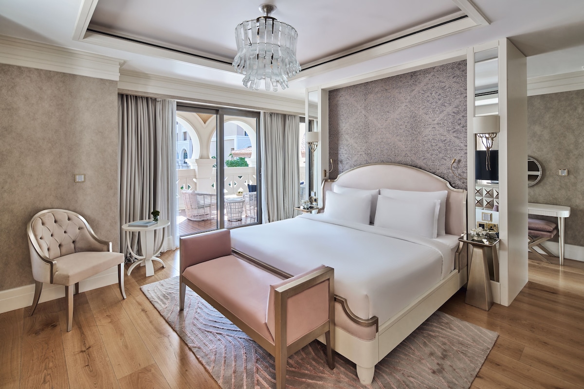 A spacious bedroom with a large bed, a seating area, a small table, and a view of the outdoors.