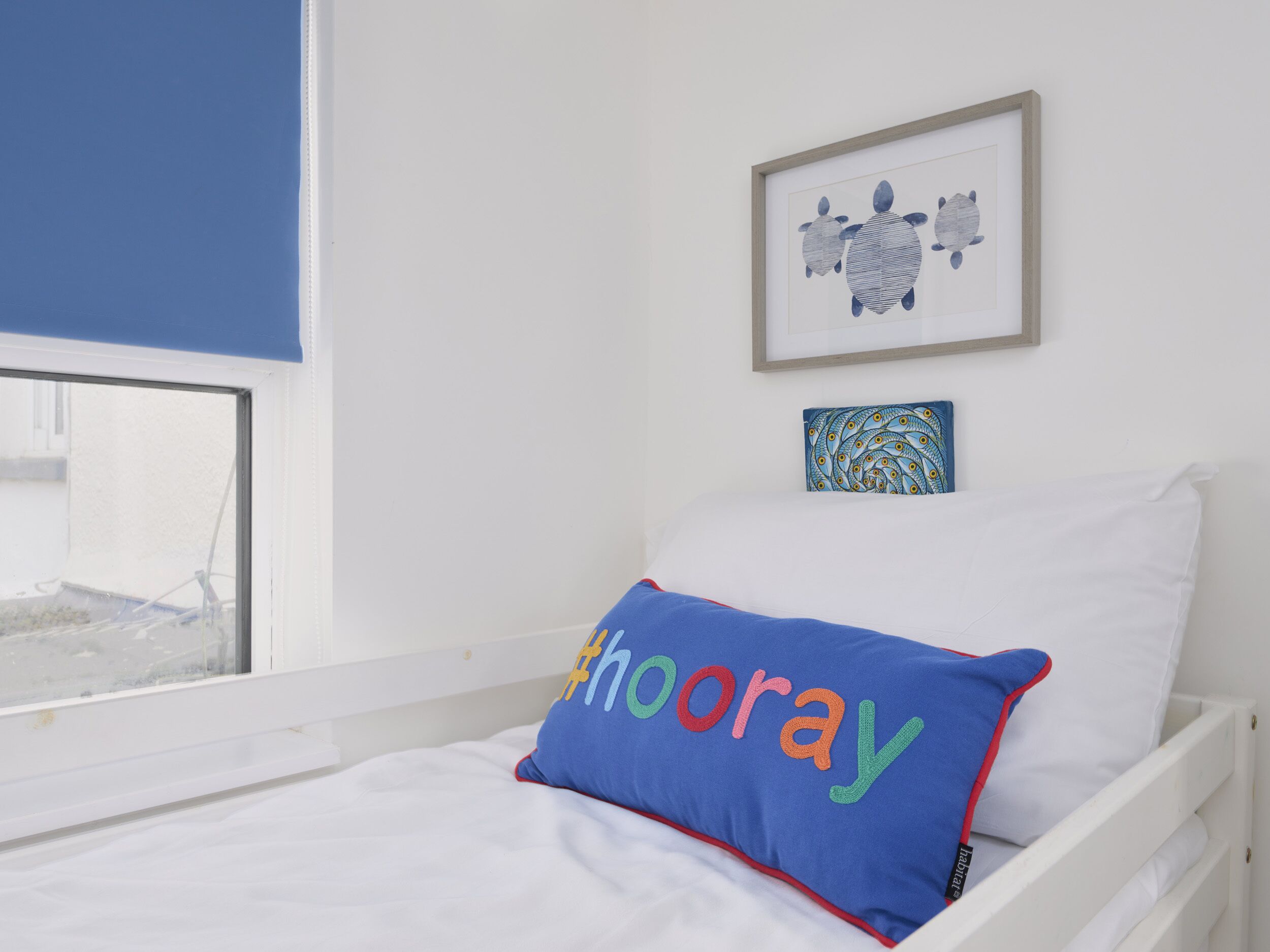A bedroom with a white bed, a blue pillow with multicolored text, a framed picture of turtles, and a window with a view of a building.