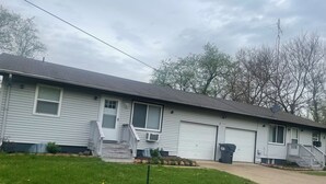 A single-story building with a garage, a covered porch, and a small front yard with grass and a few dandelions.