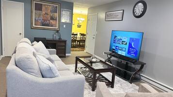 A living room with a grey sofa, a coffee table, a flat-screen TV, and a wall clock.