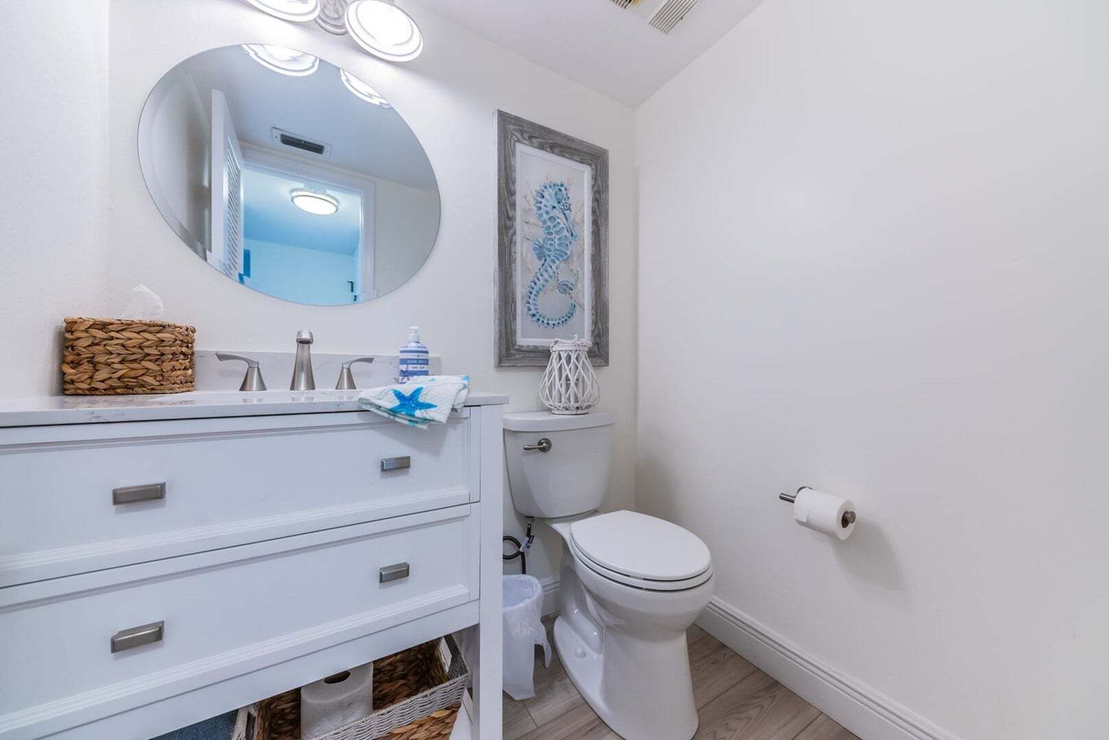 A bathroom with a white vanity, a round mirror with lights, a toilet, and a framed artwork on the wall.