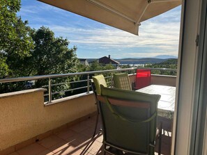 A balcony with a table and chairs, a red trash bin, and a view of trees and distant mountains. - Holiday apartment with pool and balcony (Jadranovo)