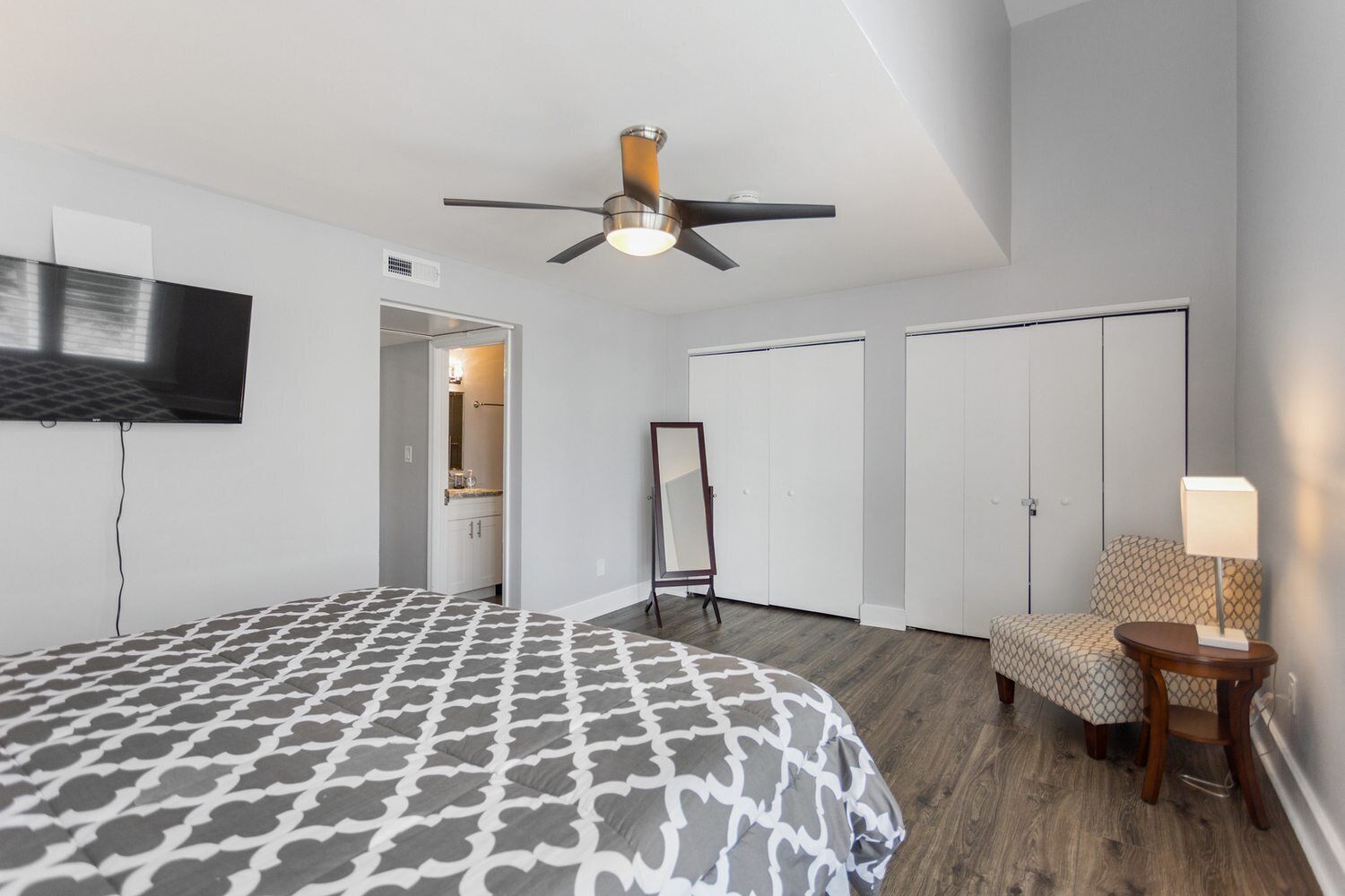 A hotel room with a bed, a ceiling fan, a TV, a mirror, a wardrobe, and a bedside table with a lamp.