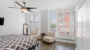 Une chambre à coucher avec un lit, deux chaises, un ventilateur de plafond et de grandes fenêtres équipées de stores.