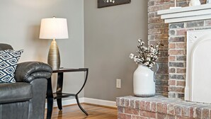 A living room with a grey sofa, a black coffee table, a white vase with flowers, and a brick fireplace.