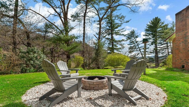 A circular seating area with Adirondack chairs around a central fire pit, set on a gravel circle in a grassy outdoor space with trees and a building in the background.