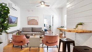 Un soggiorno moderno con un divano grigio, due poltrone rosa, un tavolino da caffè e una ventola a soffitto. C'è una TV montata a parete, un quadro incorniciato e un bancone bar in legno con una pianta.