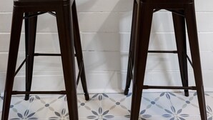 Una parete bianca con uno scaffale di legno che contiene una piccola pianta. Due sgabelli neri sono posizionati di fronte allo scaffale.