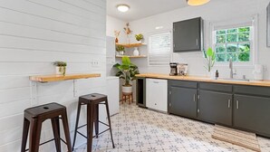 Una cucina moderna con pareti bianche, mobili scuri e un piano di lavoro in legno. Si notano due sgabelli, una pianta in vaso e una macchina per il caffè sul bancone.