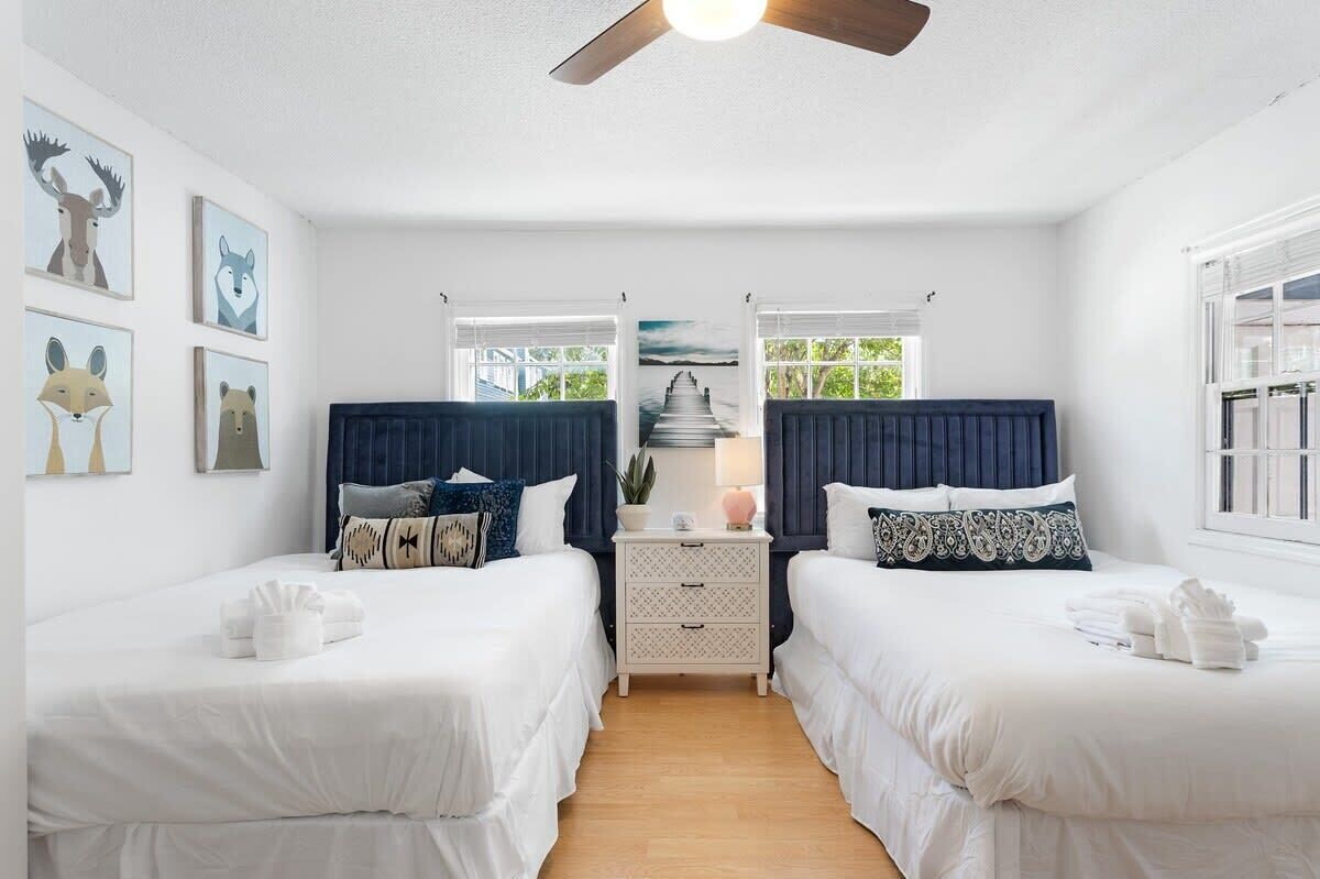 A hotel room with two beds, a ceiling fan, a nightstand with a lamp, and artwork on the walls.