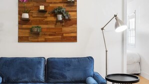 A modern living room with a blue sofa, a wooden wall-mounted shelf with plants, a round table with a bowl, and a floor lamp. - Heart of Downtown - 1 Bedroom Suite w/ Parking! (Charleston)
