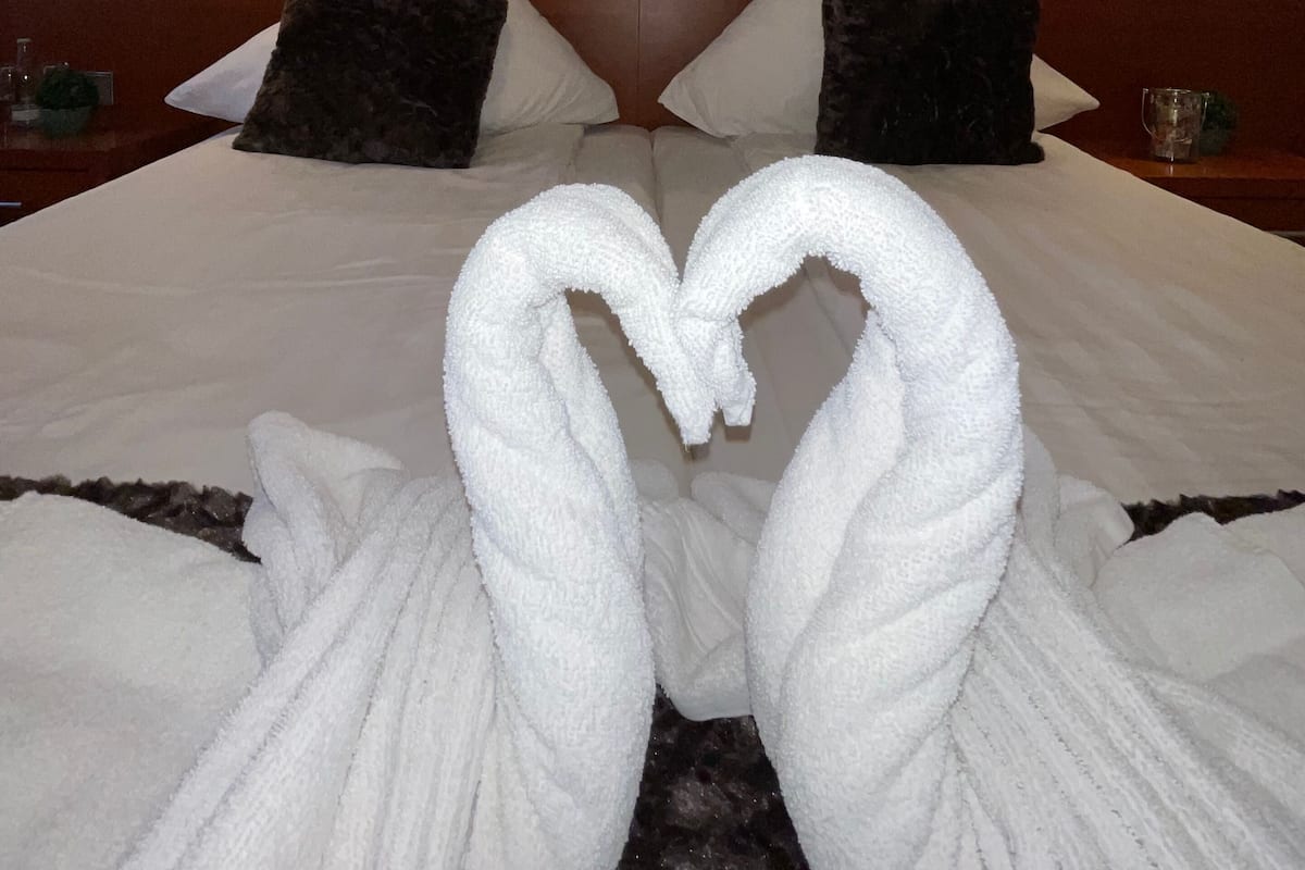 A hotel room with a neatly made bed, two pillows, a headboard, bedside lamps, and a heart-shaped towel decoration.