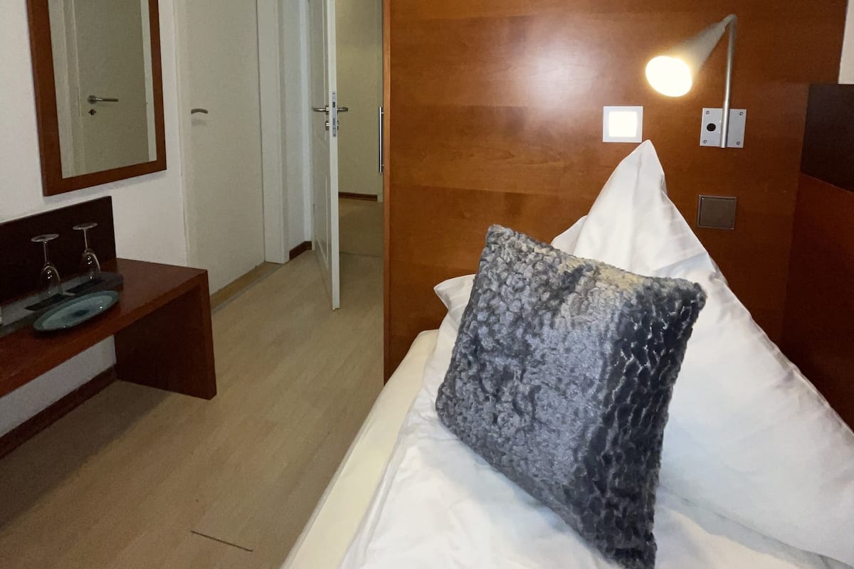 A hotel room with a wooden headboard, a bedside lamp, a mirror, and a small wooden bench with a sink.