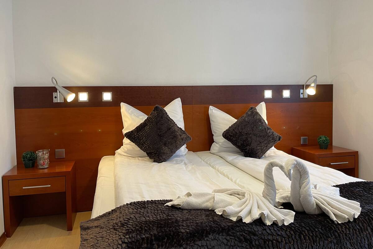 A neatly made bed with white linens, two dark pillows, and a decorative swan towel arrangement. There are two bedside tables with lamps, a small plant, and a decorative item on one table.