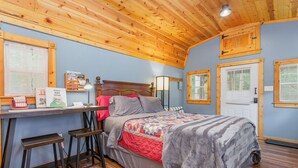 A cozy bedroom with a wooden ceiling, a bed with a quilt, a desk with a lamp, and a window. - Acorn Cottage | Adults-Only · Private Hot Tub · Secluded Hocking Hills Cabin (Logan)