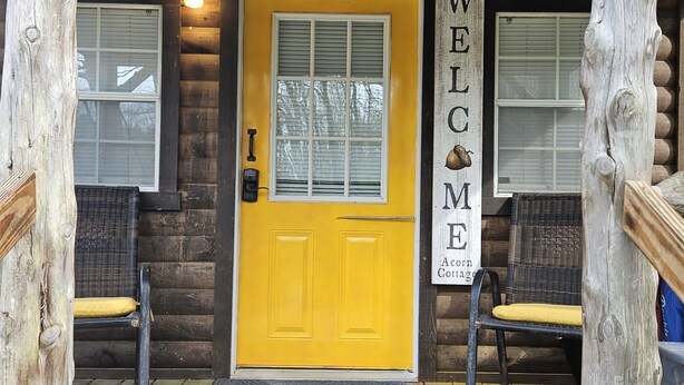 A wooden cabin with a bright yellow door, a welcome sign, and two wicker chairs outside. - Acorn Cottage | Adults-Only · Private Hot Tub · Secluded Hocking Hills Cabin (Logan)