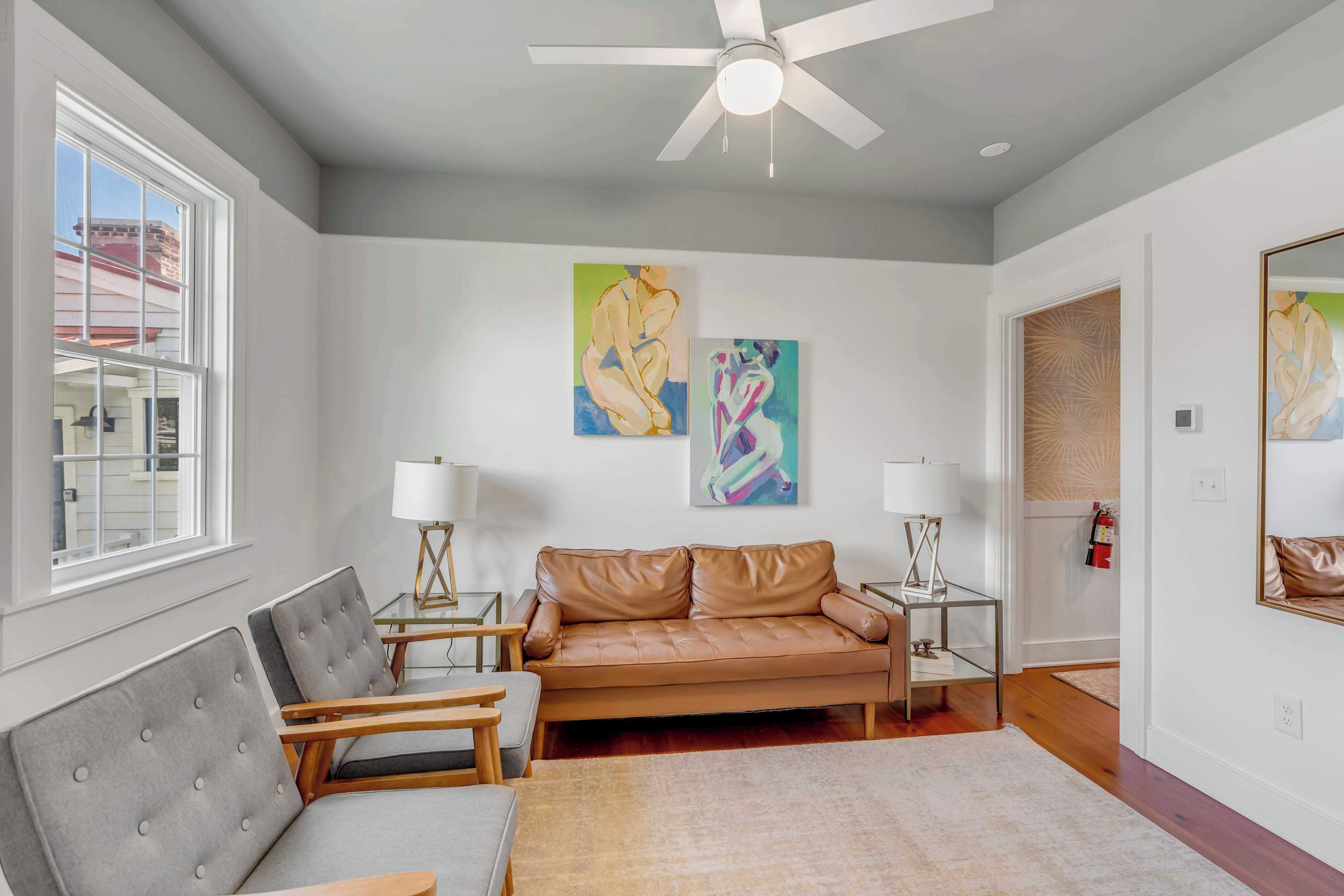 A modern living room with a brown leather sofa, two grey tufted chairs, a glass coffee table, and a ceiling fan. There are two wall-mounted lamps and abstract paintings above the sofa.