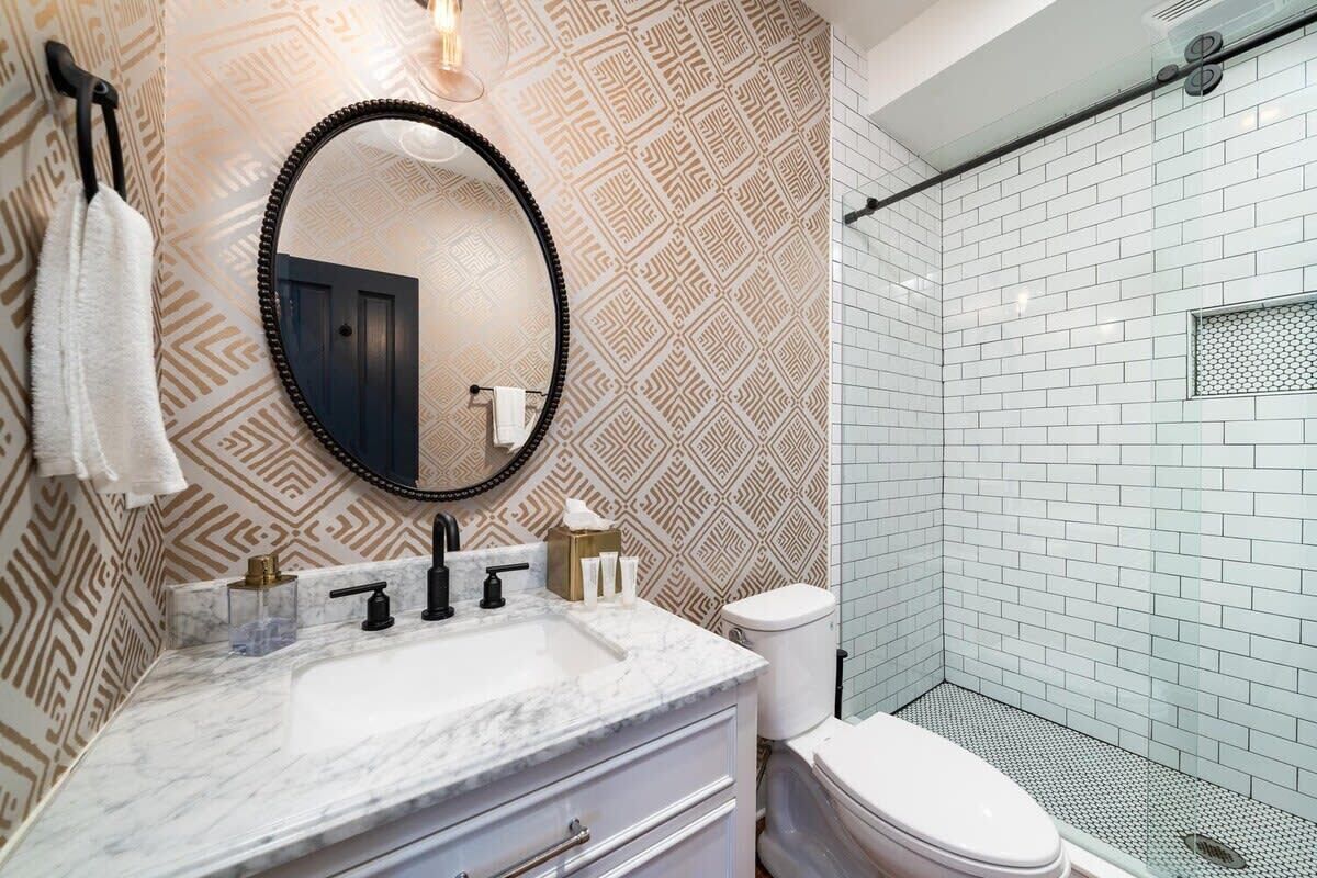 A bathroom with a patterned wall, a round mirror, a marble sink, a toilet, and a glass shower enclosure.