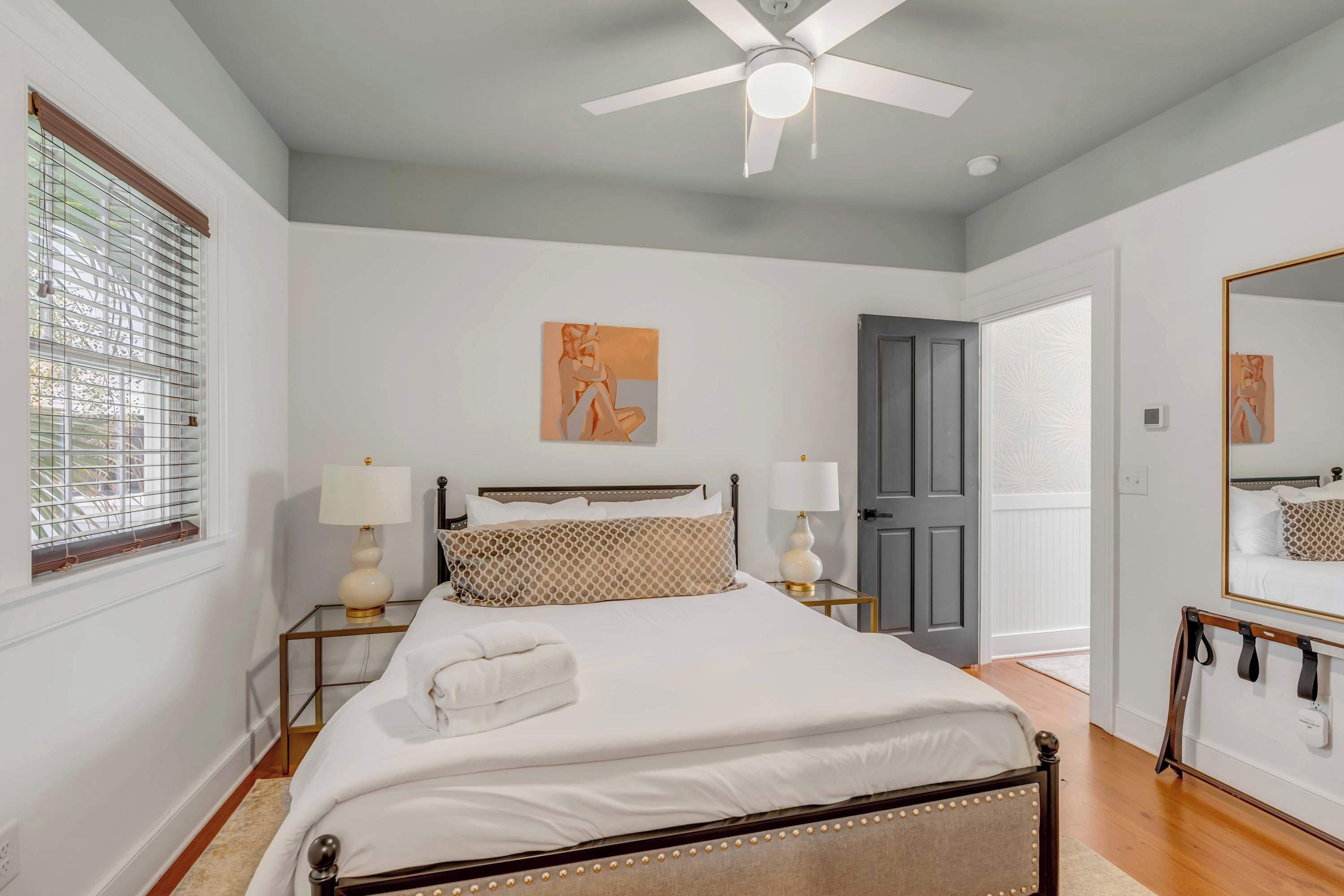Une chambre à coucher avec un lit, deux tables de chevet avec des lampes, un ventilateur de plafond, une porte et un grand miroir.